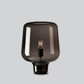 A Clérachic smoke grey glass table lamp on a light grey backdrop, its curved shade flaring over a short glass foot.
