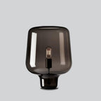 A Clérachic smoke grey glass table lamp on a light grey backdrop, its curved shade flaring over a short glass foot.