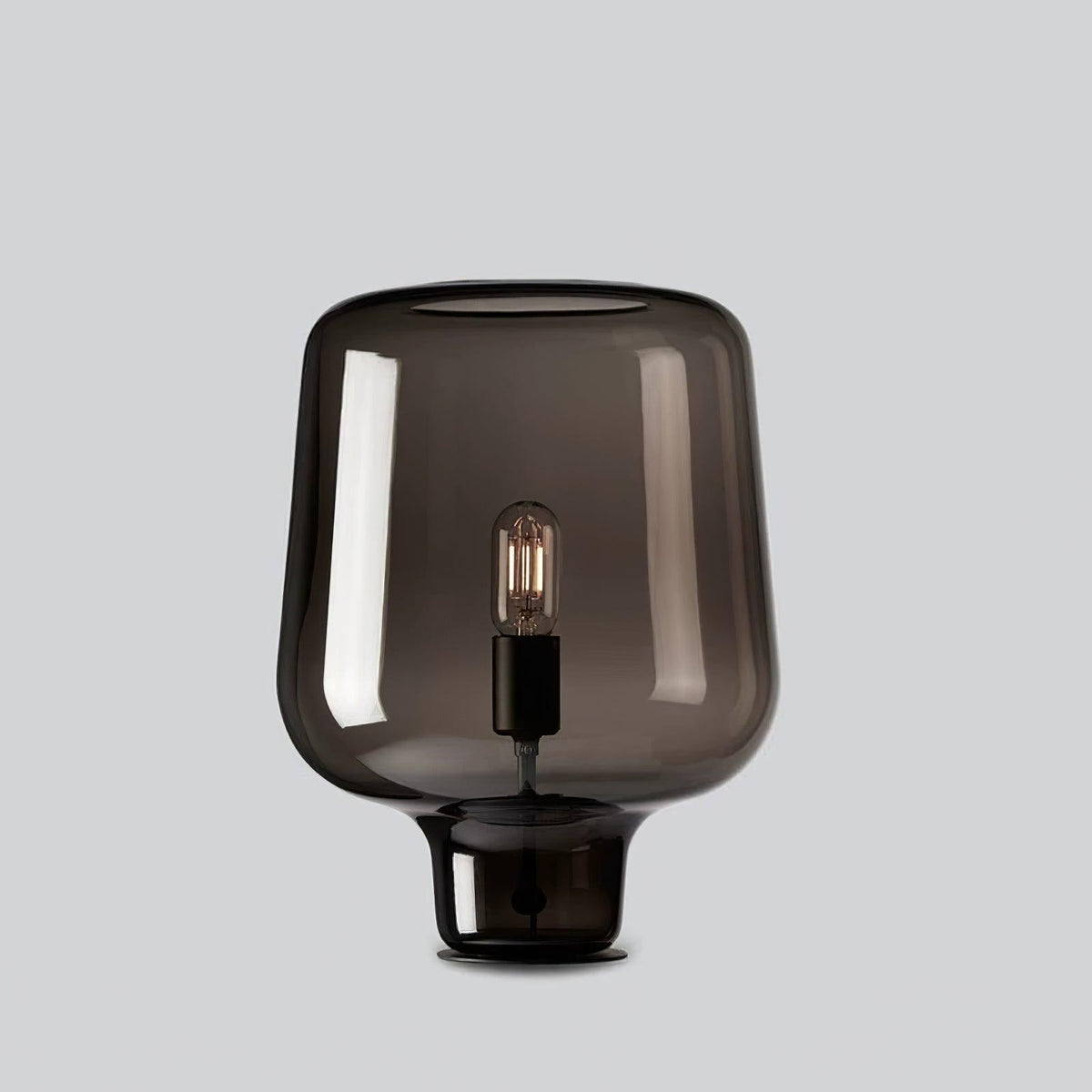 A Clérachic smoke grey glass table lamp on a light grey backdrop, its curved shade flaring over a short glass foot.
