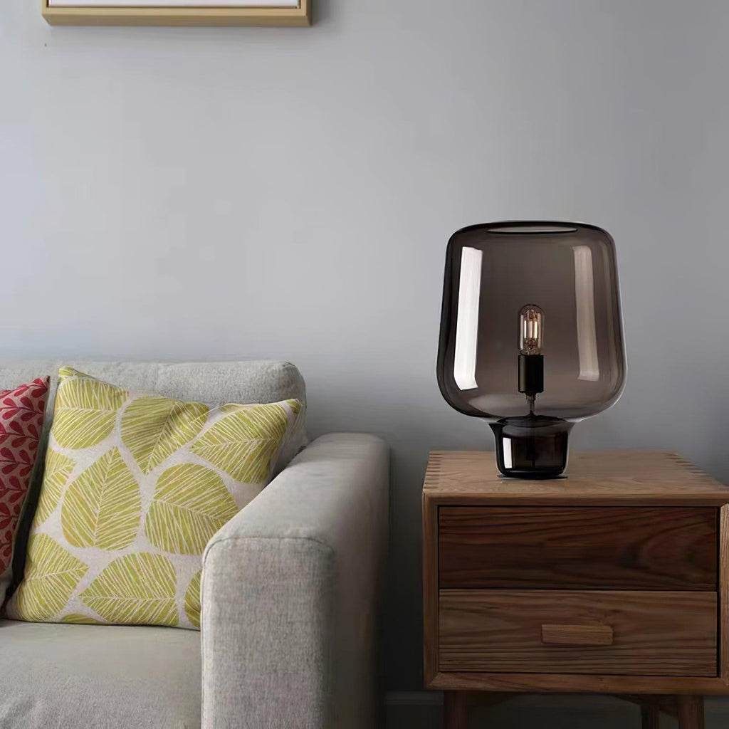 This Clérachic glass table lamp sits on a wooden nightstand beside a sofa with a yellow leaf-print cushion, against a grey wall.