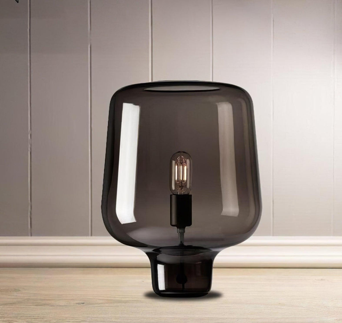 A Clérachic table lamp on a pale wood floor in front of a panelled wall, its smoke grey glass shade filling the frame.