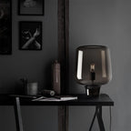 On a black desk in a dark grey room, this Clérachic glass table lamp stands near a stoneware bottle and a framed print.