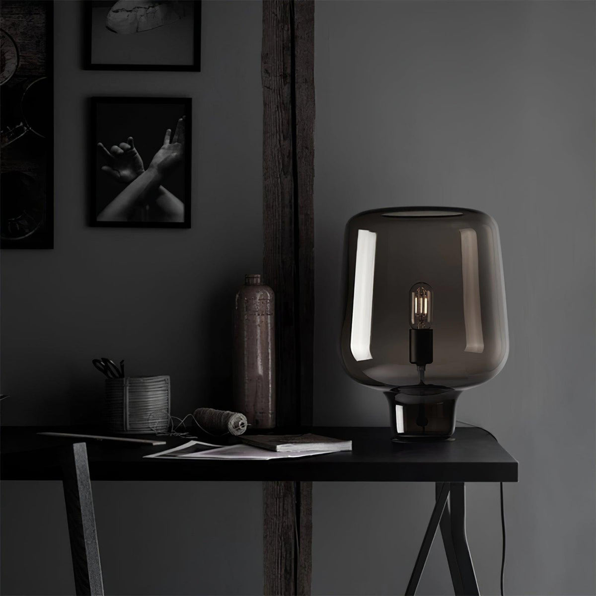 On a black desk in a dark grey room, this Clérachic glass table lamp stands near a stoneware bottle and a framed print.