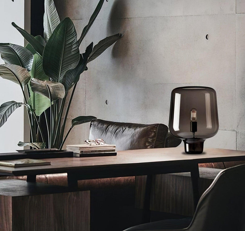 Beside a large leafy plant and a leather sofa, a Clérachic smoke grey glass lamp stands on a wooden desk.
