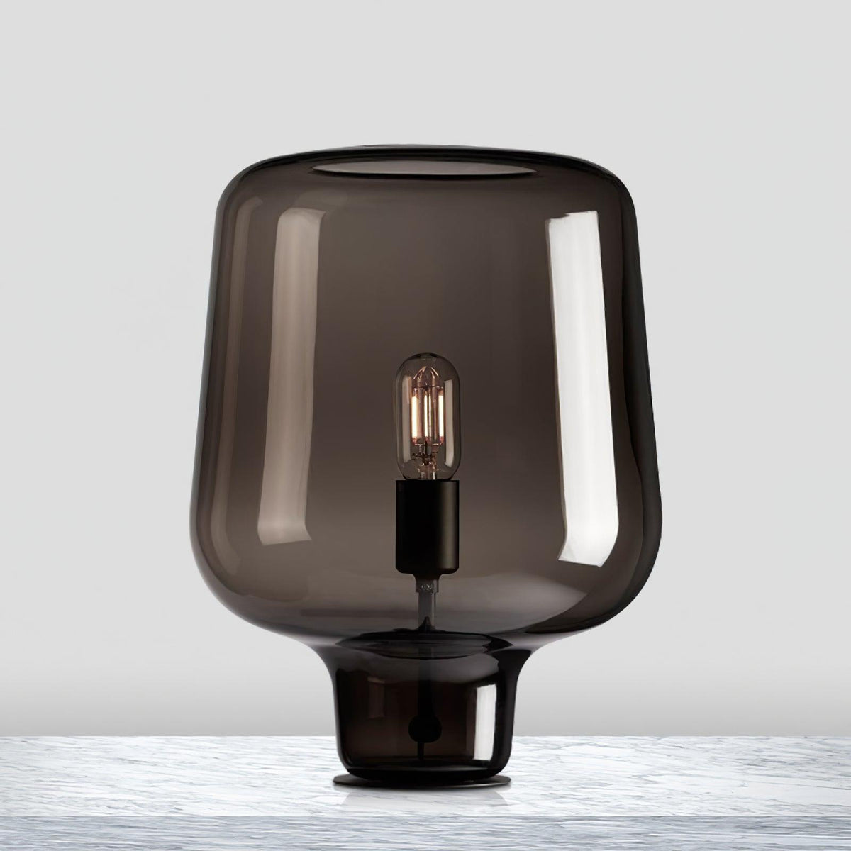 Close view of this Clérachic smoke grey glass table lamp on a pale marble surface, light grey backdrop behind.