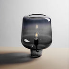 A Clérachic glass table lamp in a darker blue-grey tint on a pale tabletop, its filament bulb faintly visible inside.