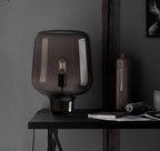 On a black desk with a stoneware bottle, twine and a book, this Clérachic glass table lamp stands against a dark wall.