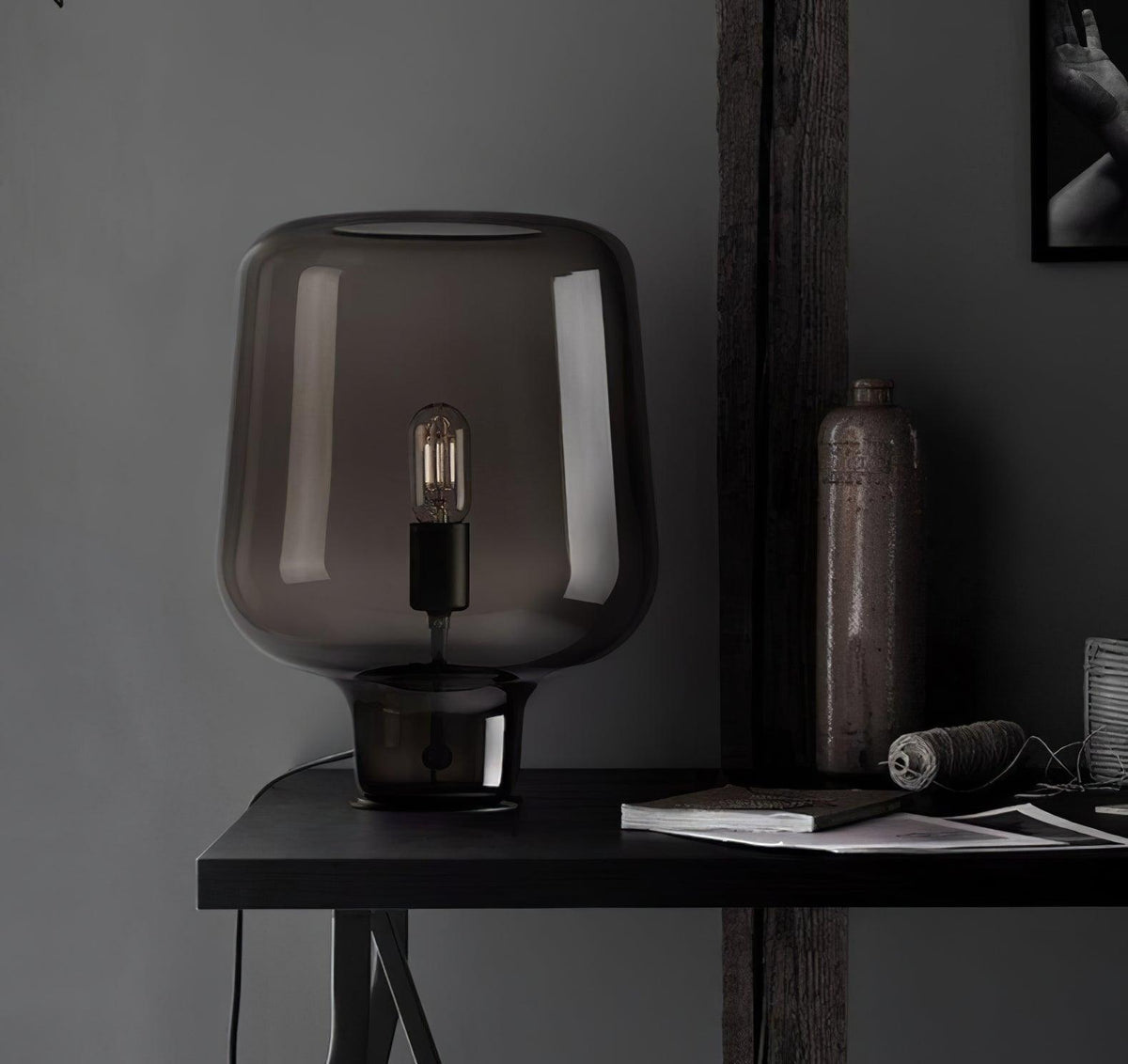 On a black desk with a stoneware bottle, twine and a book, this Clérachic glass table lamp stands against a dark wall.