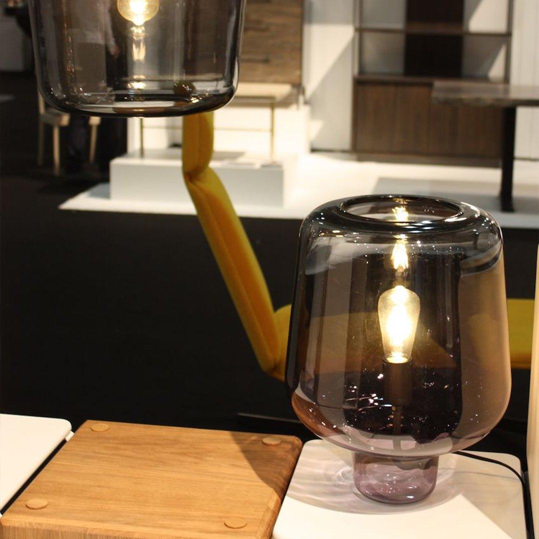 At an exhibition, a Clérachic glass table lamp glows on a pale wood stool, another glass shade hanging above it.