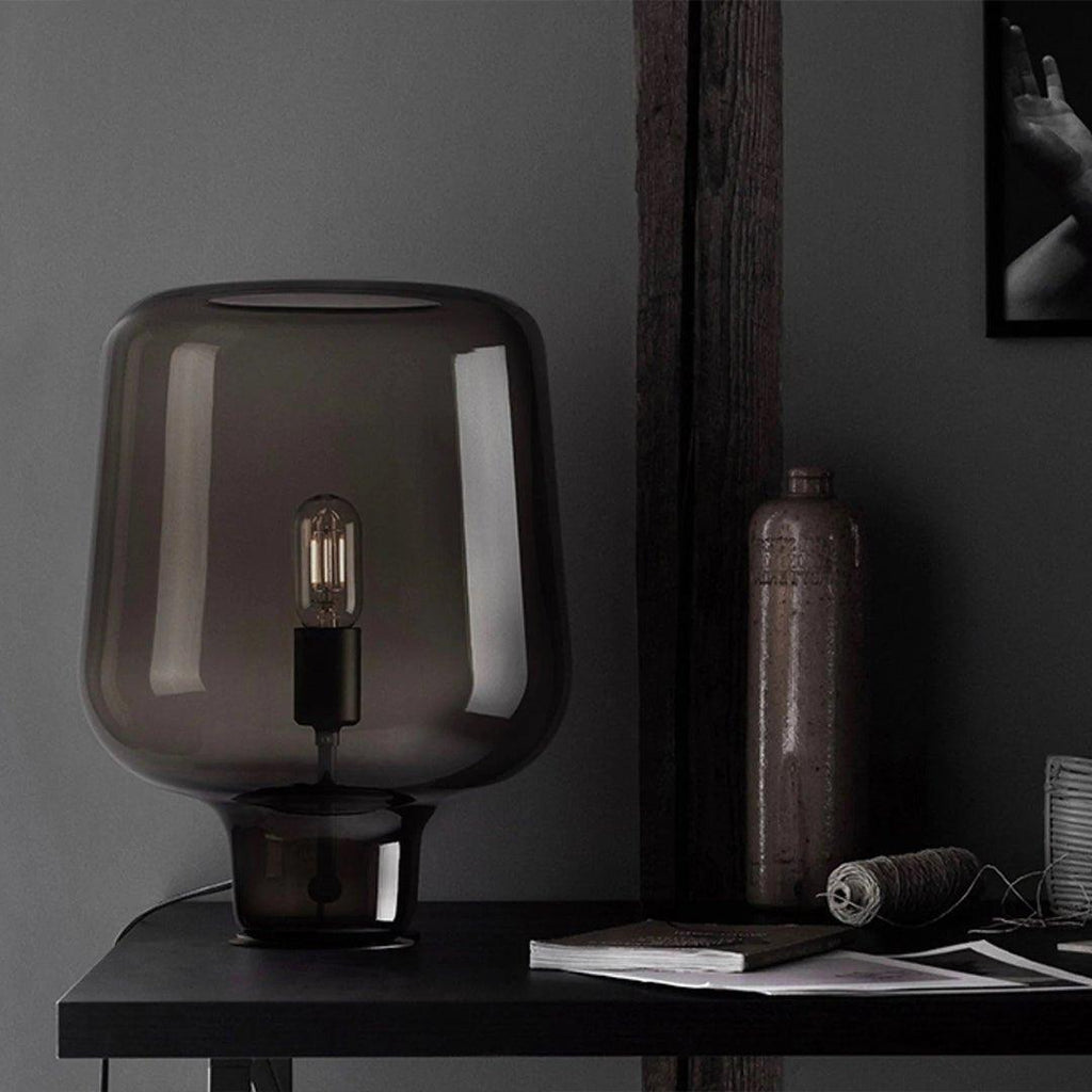 Close view of this Clérachic smoke grey glass table lamp on a dark desk, beside a bottle, a spool of twine and a book.