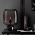 Close view of this Clérachic smoke grey glass table lamp on a dark desk, beside a bottle, a spool of twine and a book.