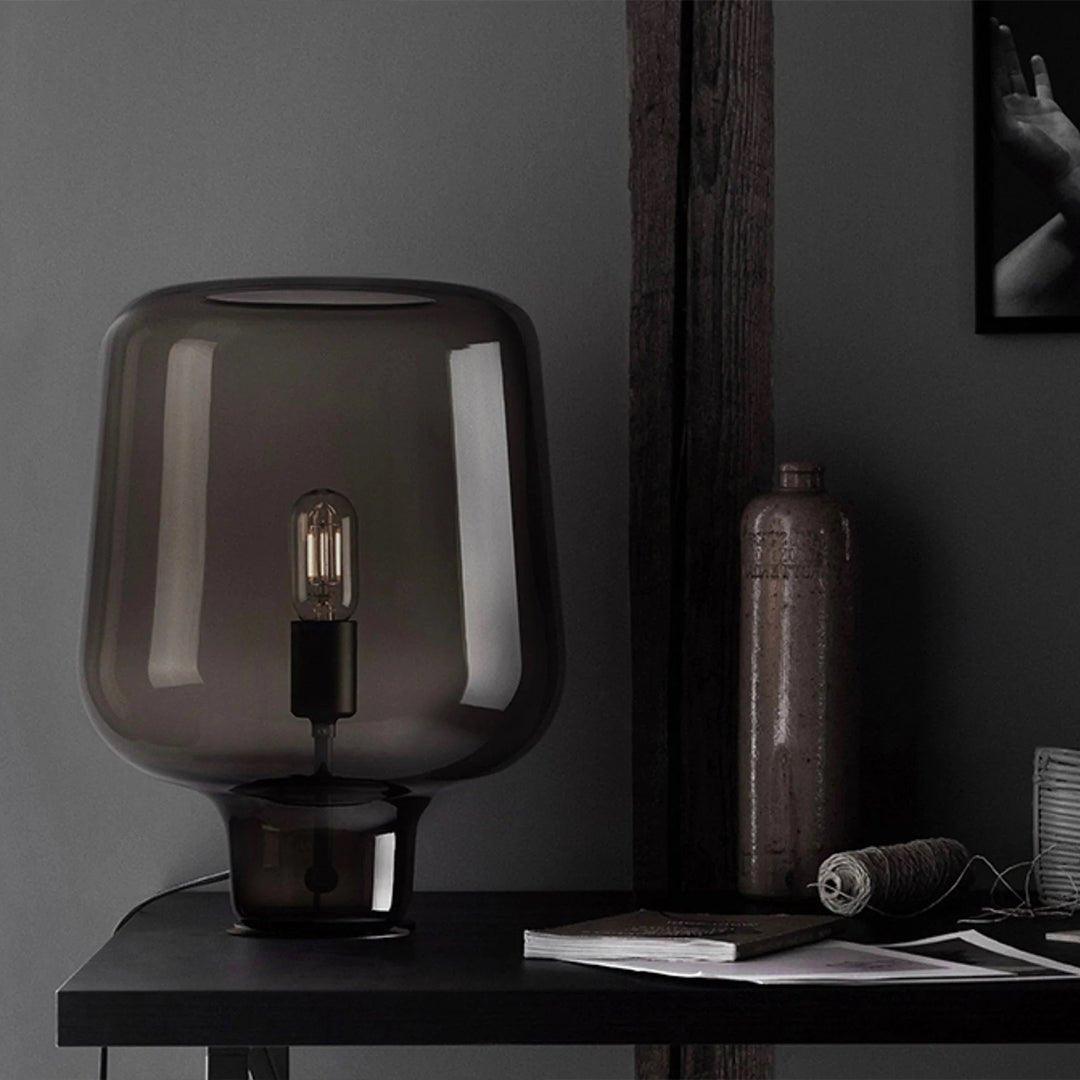 Close view of this Clérachic smoke grey glass table lamp on a dark desk, beside a bottle, a spool of twine and a book.