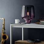 A plum-tinted glass table lamp from Clérachic on a white desk, with a mug, a synthesiser and a saxophone beside it.