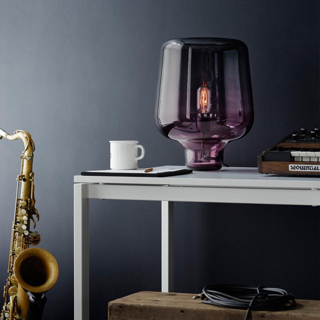 A plum-tinted glass table lamp from Clérachic on a white desk, with a mug, a synthesiser and a saxophone beside it.