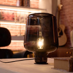 Lit Clérachic glass table lamp on a pale wood table in a warm brick-walled room, a guitar blurred behind it.