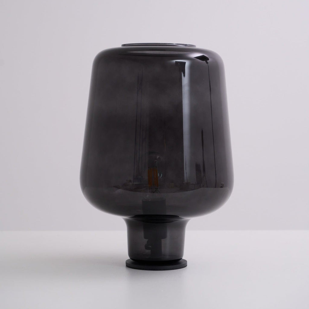Unlit, this Clérachic smoke grey glass table lamp rests on a white surface against a soft grey-lilac backdrop.