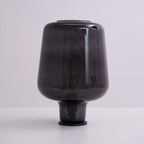 Unlit, this Clérachic smoke grey glass table lamp rests on a white surface against a soft grey-lilac backdrop.