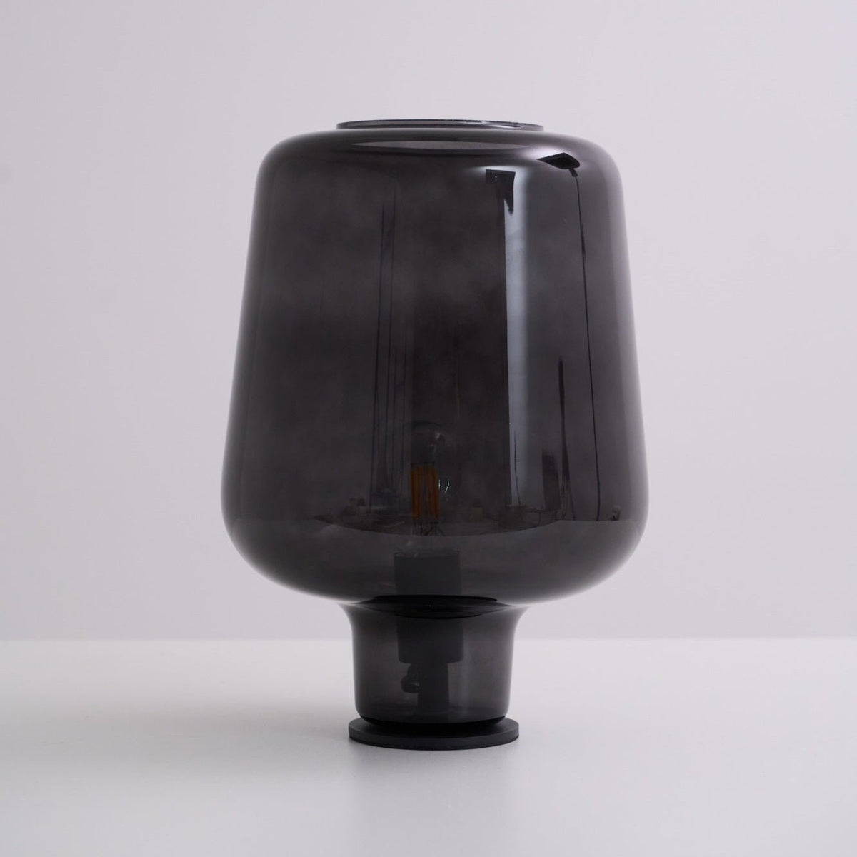 Unlit, this Clérachic smoke grey glass table lamp rests on a white surface against a soft grey-lilac backdrop.
