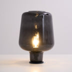 Warm light glows from the filament bulb inside this Clérachic smoke grey glass table lamp, on a white surface.