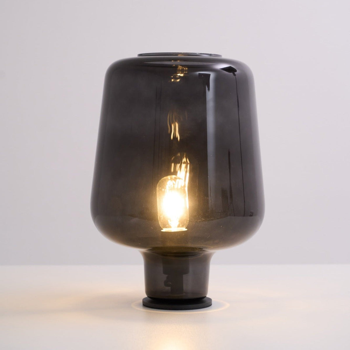 Warm light glows from the filament bulb inside this Clérachic smoke grey glass table lamp, on a white surface.