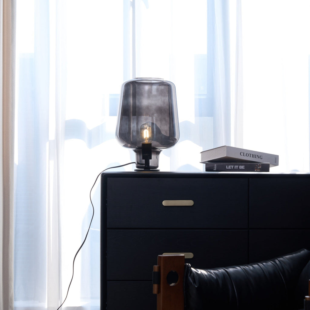 Further back, a Clérachic glass table lamp on a black chest with brass handles, a cord trailing down the side and curtains behind.
