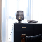 Further back, a Clérachic glass table lamp on a black chest with brass handles, a cord trailing down the side and curtains behind.