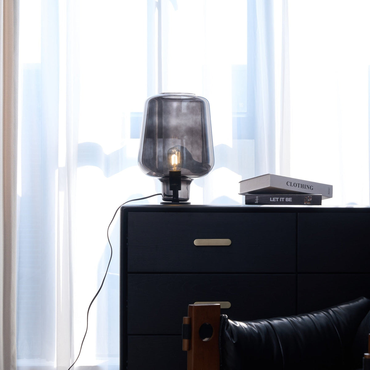 Further back, a Clérachic glass table lamp on a black chest with brass handles, a cord trailing down the side and curtains behind.