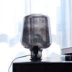 Close view of a Clérachic smoke grey glass lamp, unlit, on a black chest of drawers against bright sheer curtains.