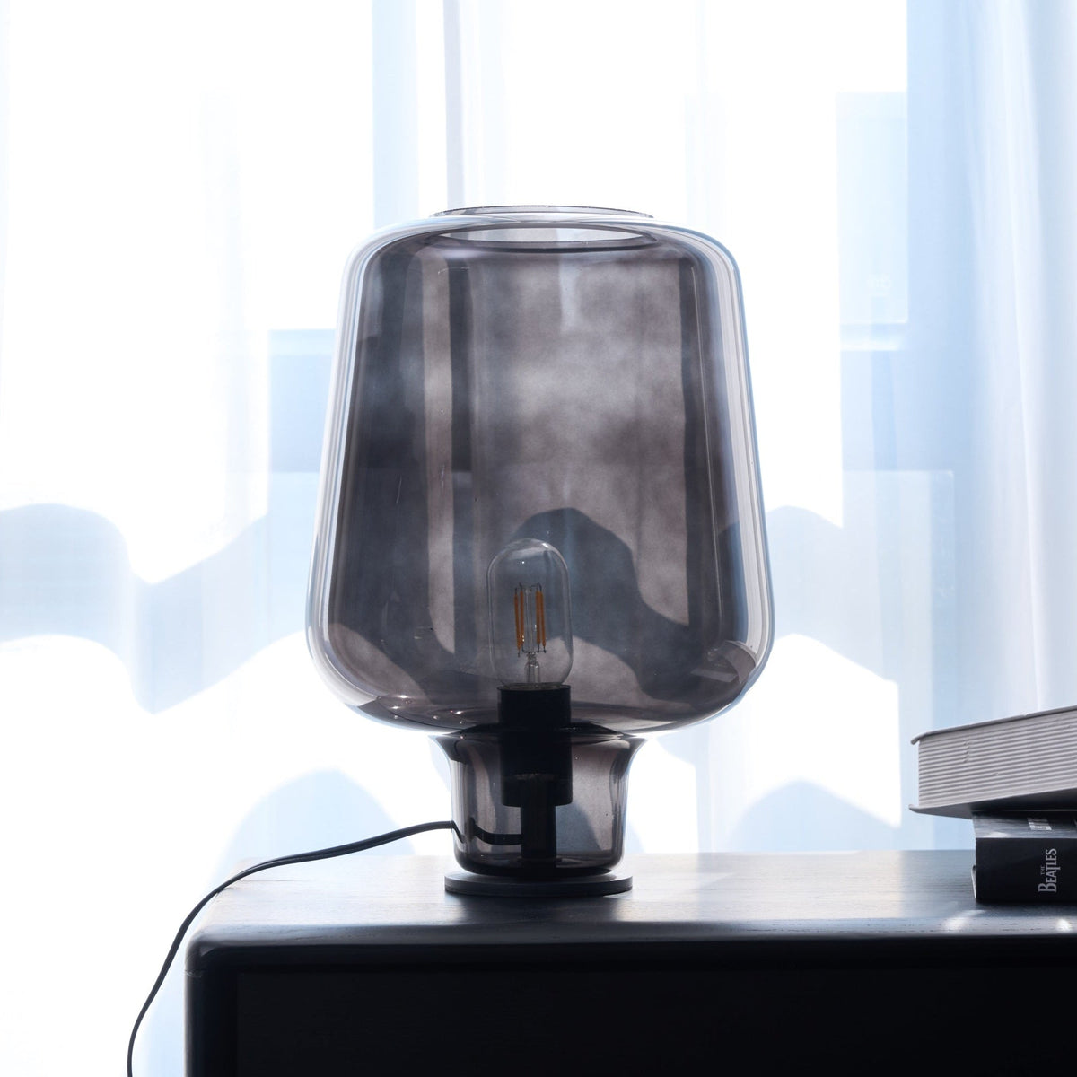 Close view of a Clérachic smoke grey glass lamp, unlit, on a black chest of drawers against bright sheer curtains.