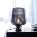 This Clérachic glass table lamp, lit, with its filament bulb glowing inside the smoke grey shade, a book stack at its side.