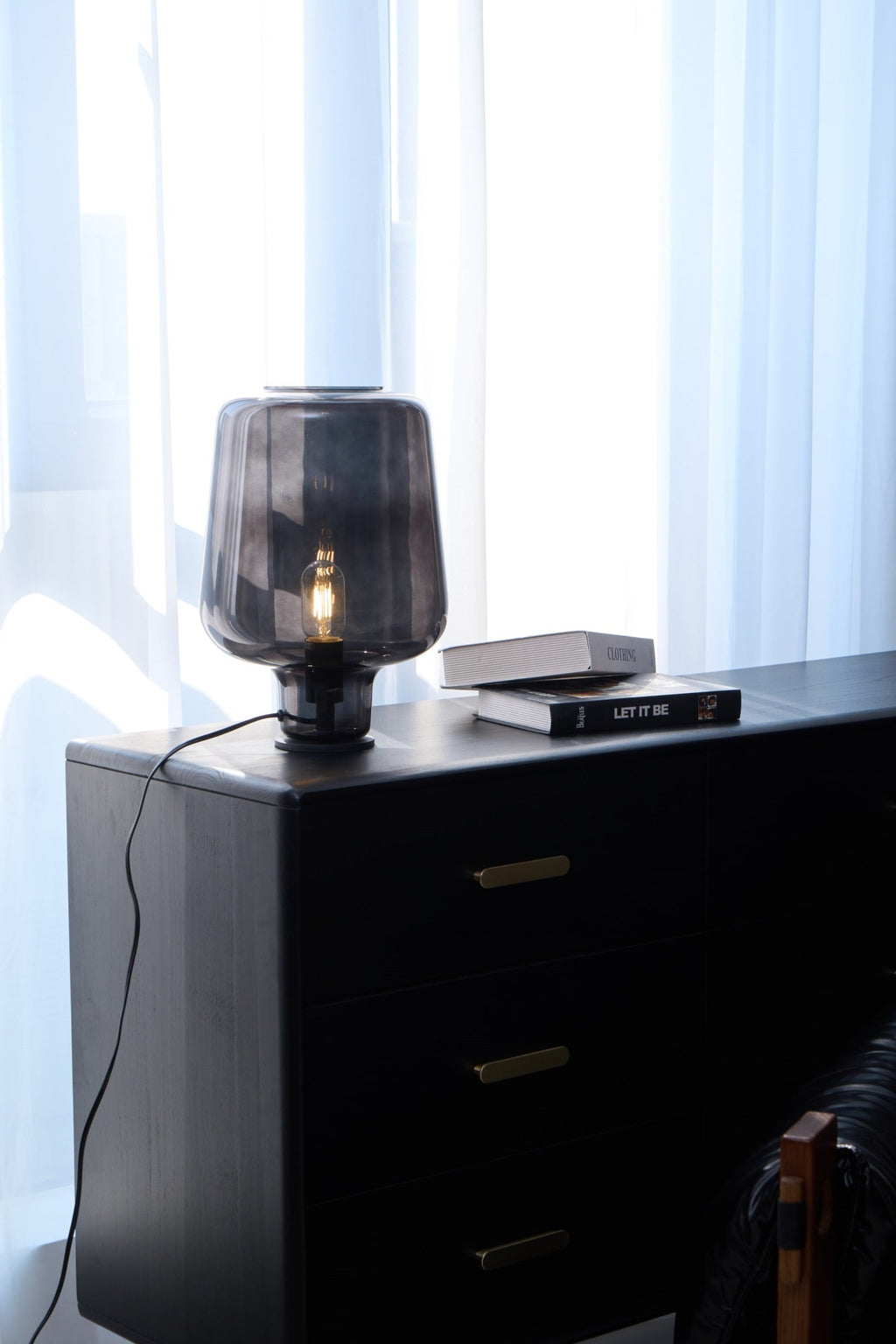 A Clérachic glass table lamp lit on a black chest of drawers, an upright shot with sheer curtains and stacked books.