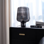 Beside beige curtains, a Clérachic smoke grey glass lamp stands on a black chest with a brass pull and two books.