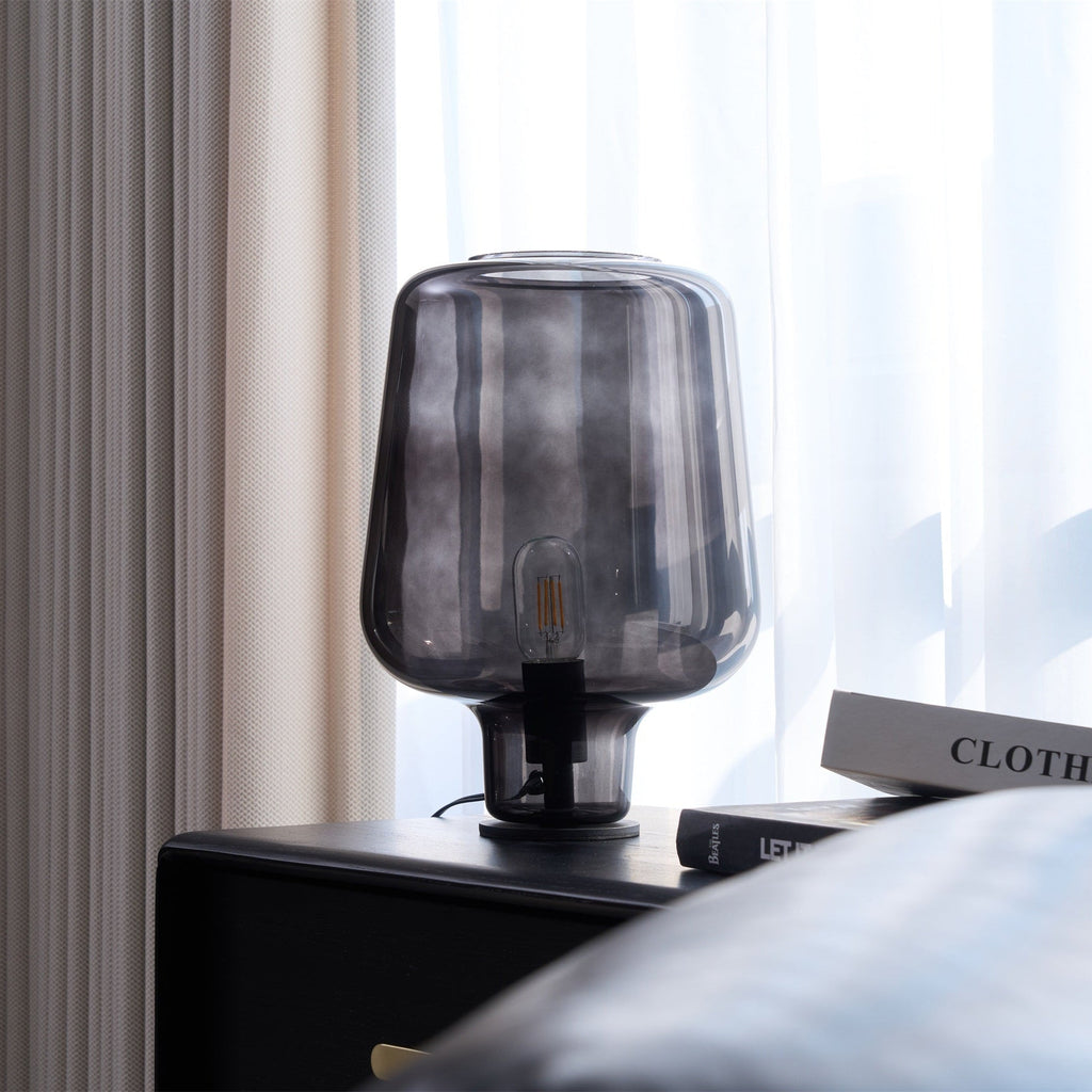 At a slight angle, this Clérachic glass table lamp stands on a black cabinet, with a leather cushion at the bottom corner.