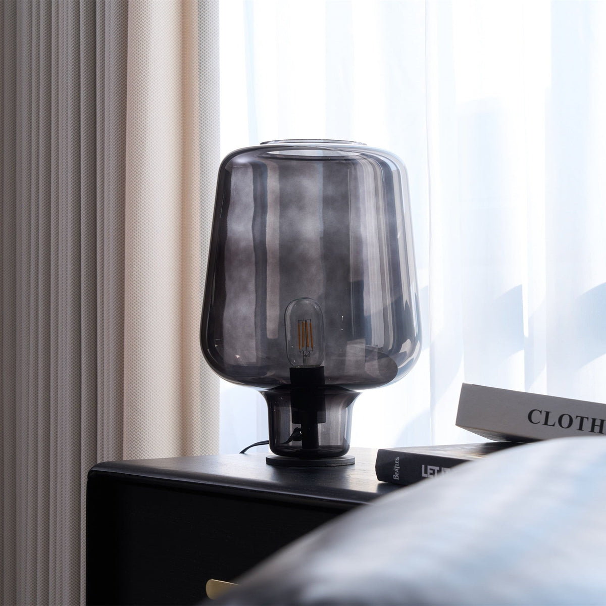 At a slight angle, this Clérachic glass table lamp stands on a black cabinet, with a leather cushion at the bottom corner.