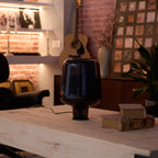 Dim evening light on an unlit Clérachic glass table lamp, with old books on a pale wood table and brick shelves behind.