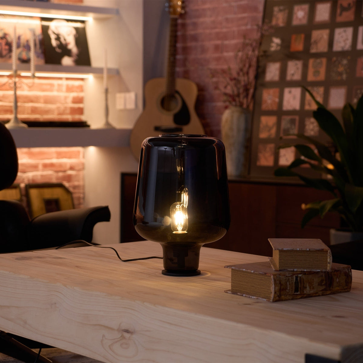 Lit Clérachic glass table lamp on a wood table, its small bulb glowing in the dusk beside a pile of old books.