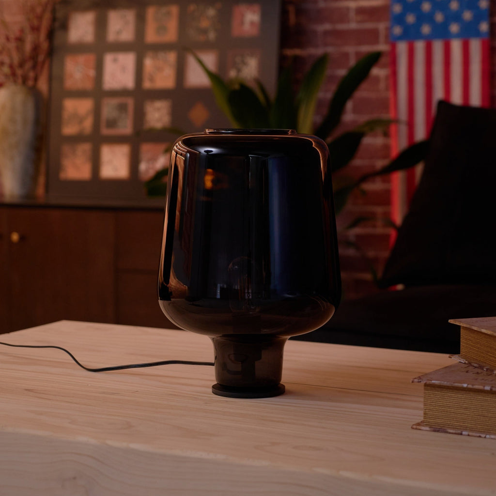 A dark, unlit Clérachic glass table lamp on a wood table, a sofa arm and a plant leaf in soft focus behind.