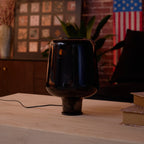 A dark, unlit Clérachic glass table lamp on a wood table, a sofa arm and a plant leaf in soft focus behind.