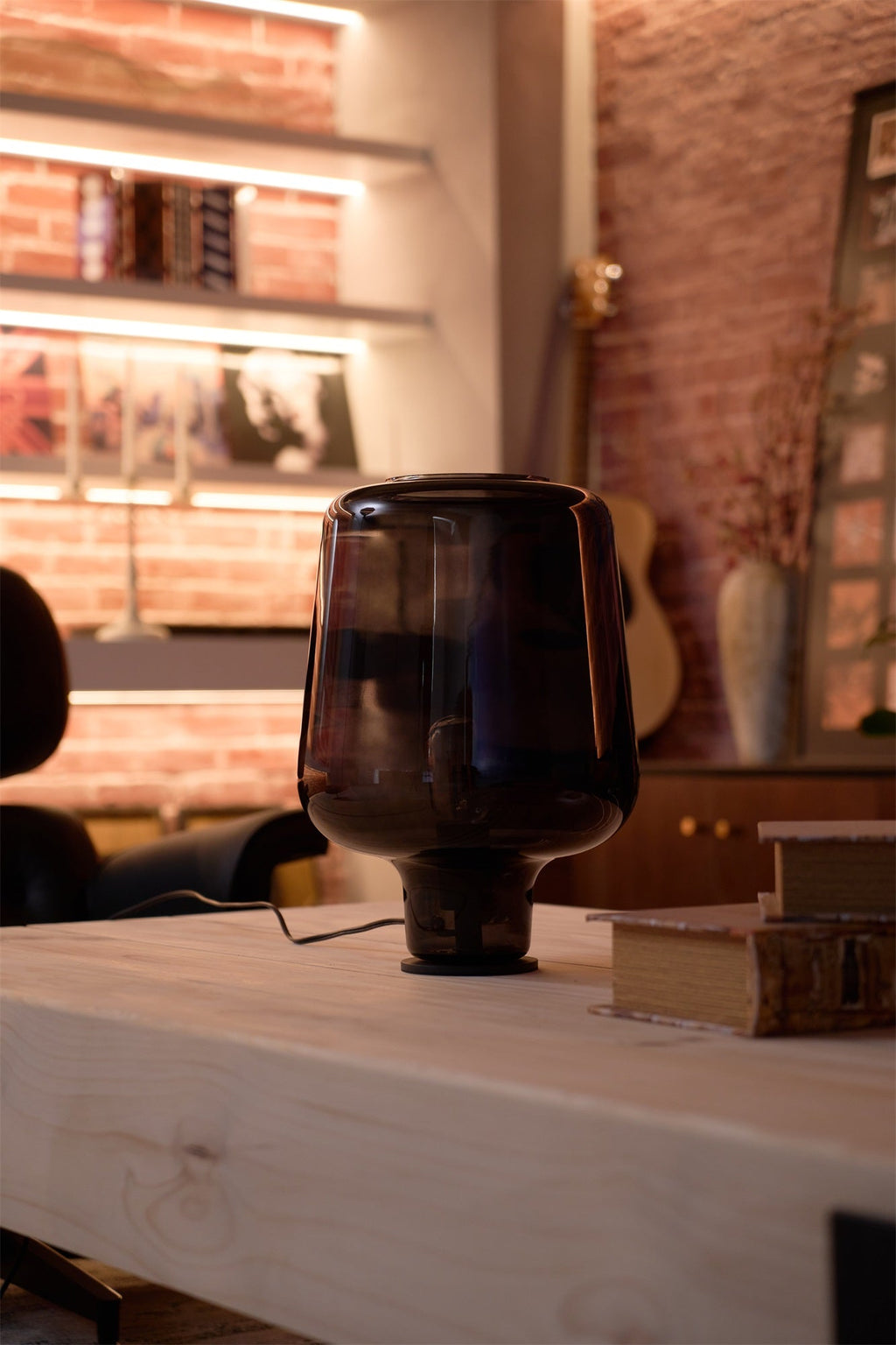 Tall view of a Clérachic glass table lamp, unlit, on a pale wood table with a stack of books in a brick-walled room.