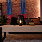A lit Clérachic glass table lamp on a pale wood coffee table, with a sofa, cushions and striped flags on a brick wall behind.