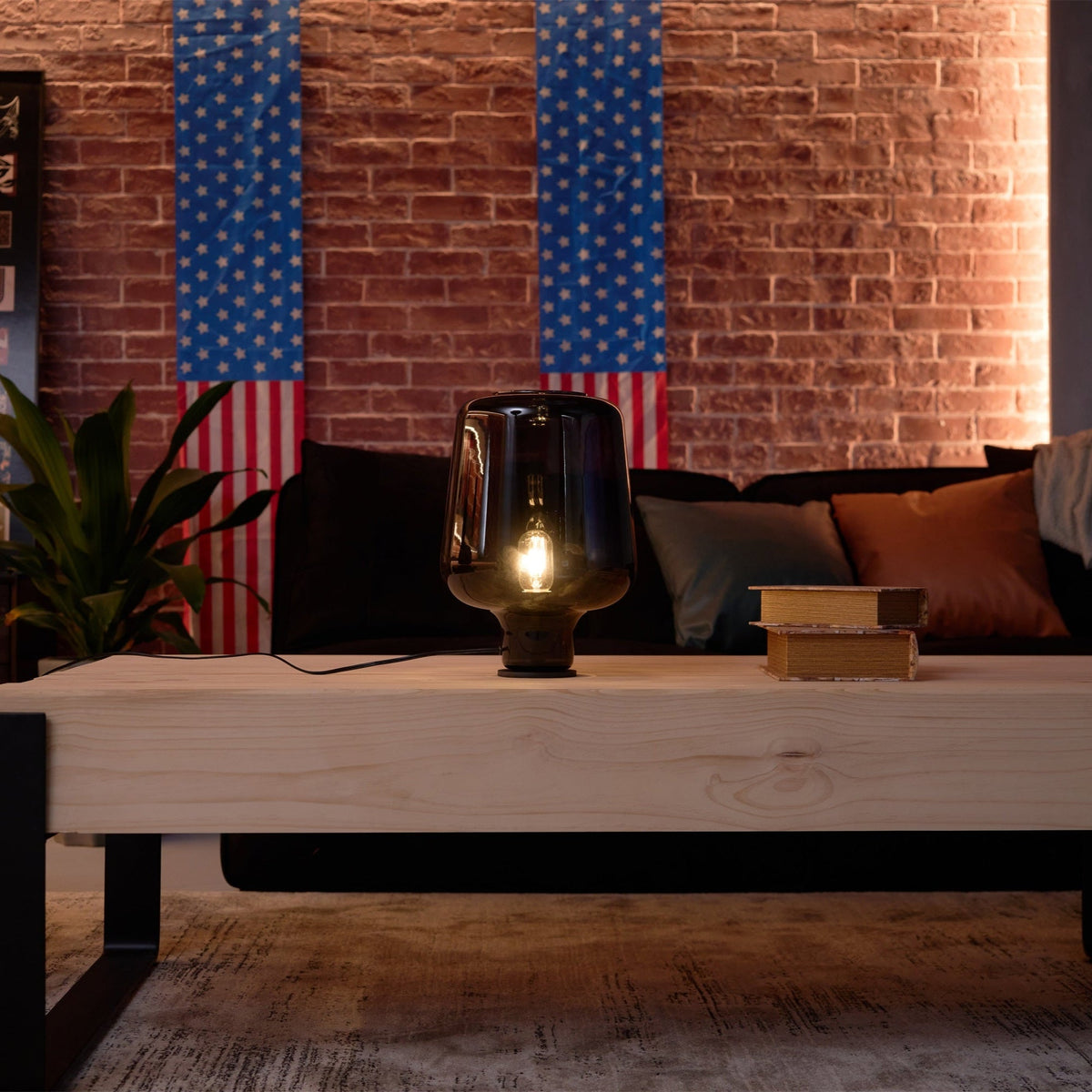 A lit Clérachic glass table lamp on a pale wood coffee table, with a sofa, cushions and striped flags on a brick wall behind.