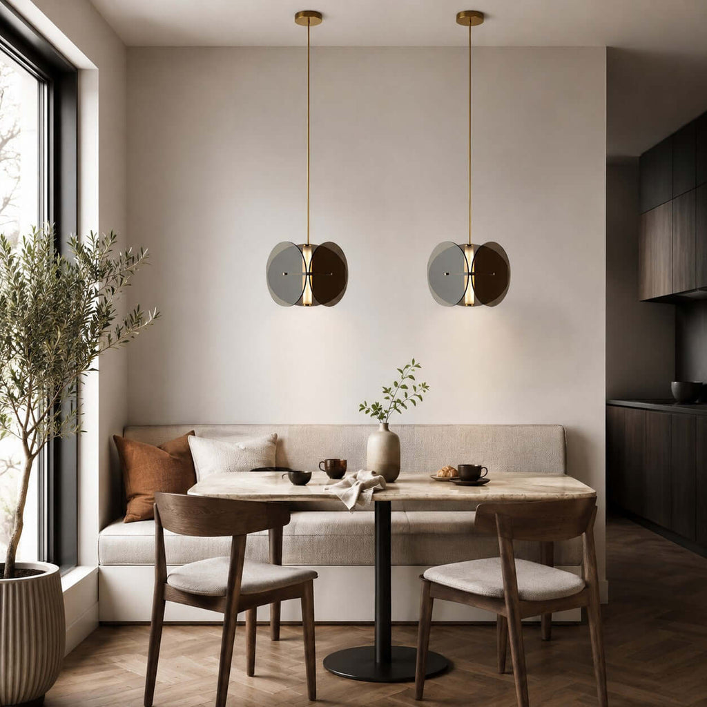 Two Clérachic disc pendants side by side above a small dining table and wooden chairs, with an olive tree by the window.