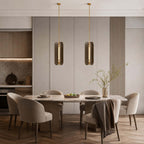 Pair of narrow Clérachic glass pendants over a long dining table with beige chairs, against a panelled grey wall.