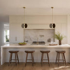 Two small Clérachic disc pendants over a cream kitchen island with four wooden saddle stools and a marble backsplash.