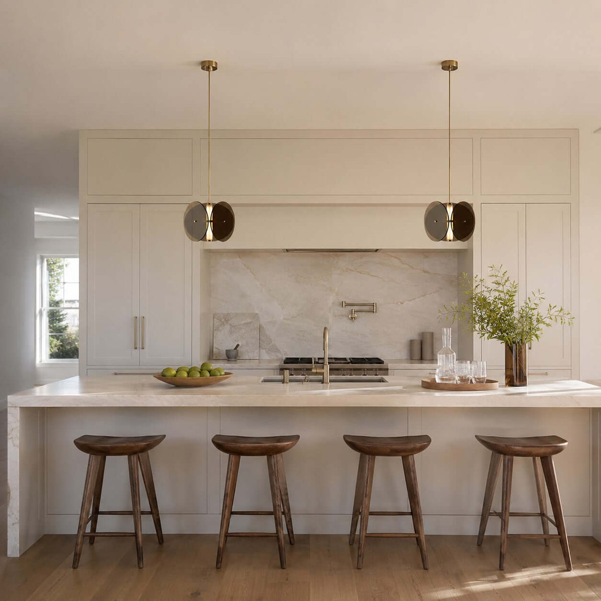 Two small Clérachic disc pendants over a cream kitchen island with four wooden saddle stools and a marble backsplash.