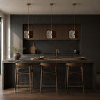 Three Clérachic smoked glass pendants above a dark kitchen island with wooden stools, against black cabinetry.