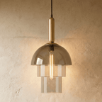 Clérachic three-tier smoked glass pendant with a brass stem and a long filament bulb glowing against a limewash wall.
