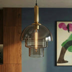 Clérachic three-tier smoked glass pendant with brass stem and tubular filament bulb, hung in front of a blue wall and walnut cabinets.