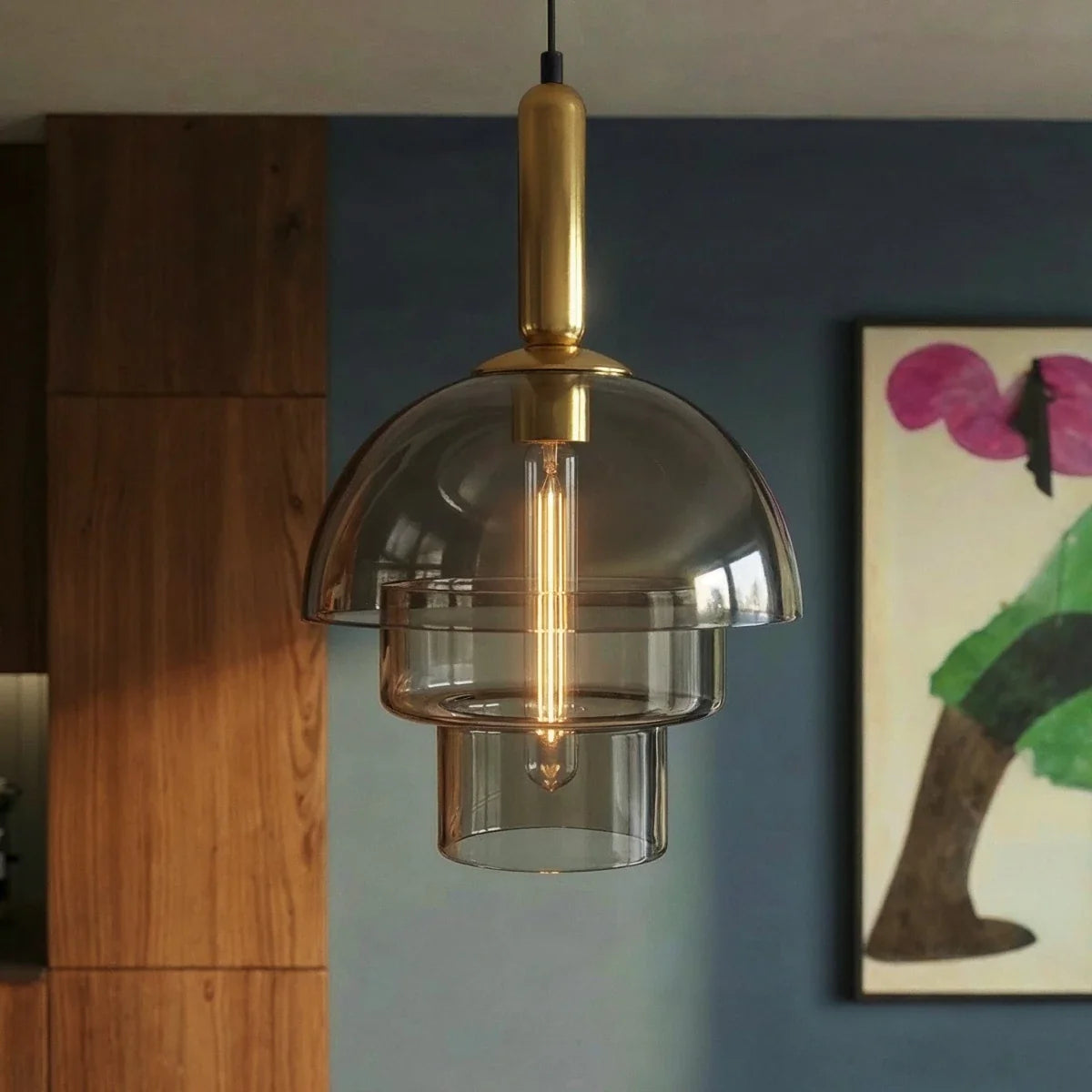 Clérachic three-tier smoked glass pendant with brass stem and tubular filament bulb, hung in front of a blue wall and walnut cabinets.
