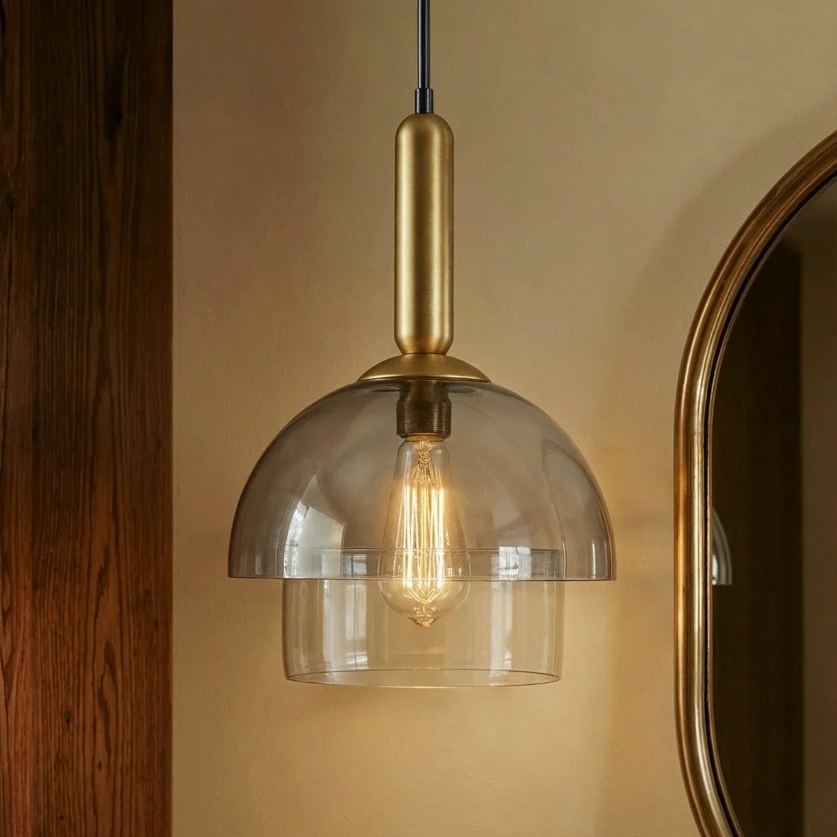 Clérachic smoked glass dome pendant with clear lower tier and brass stem, lit with a warm filament bulb beside a gold-framed mirror.